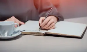 Dłoń pisze w notesie, szukając inspiracji, jak napisać intencję za zmarłego wzór. Obok filiżanka kawy.